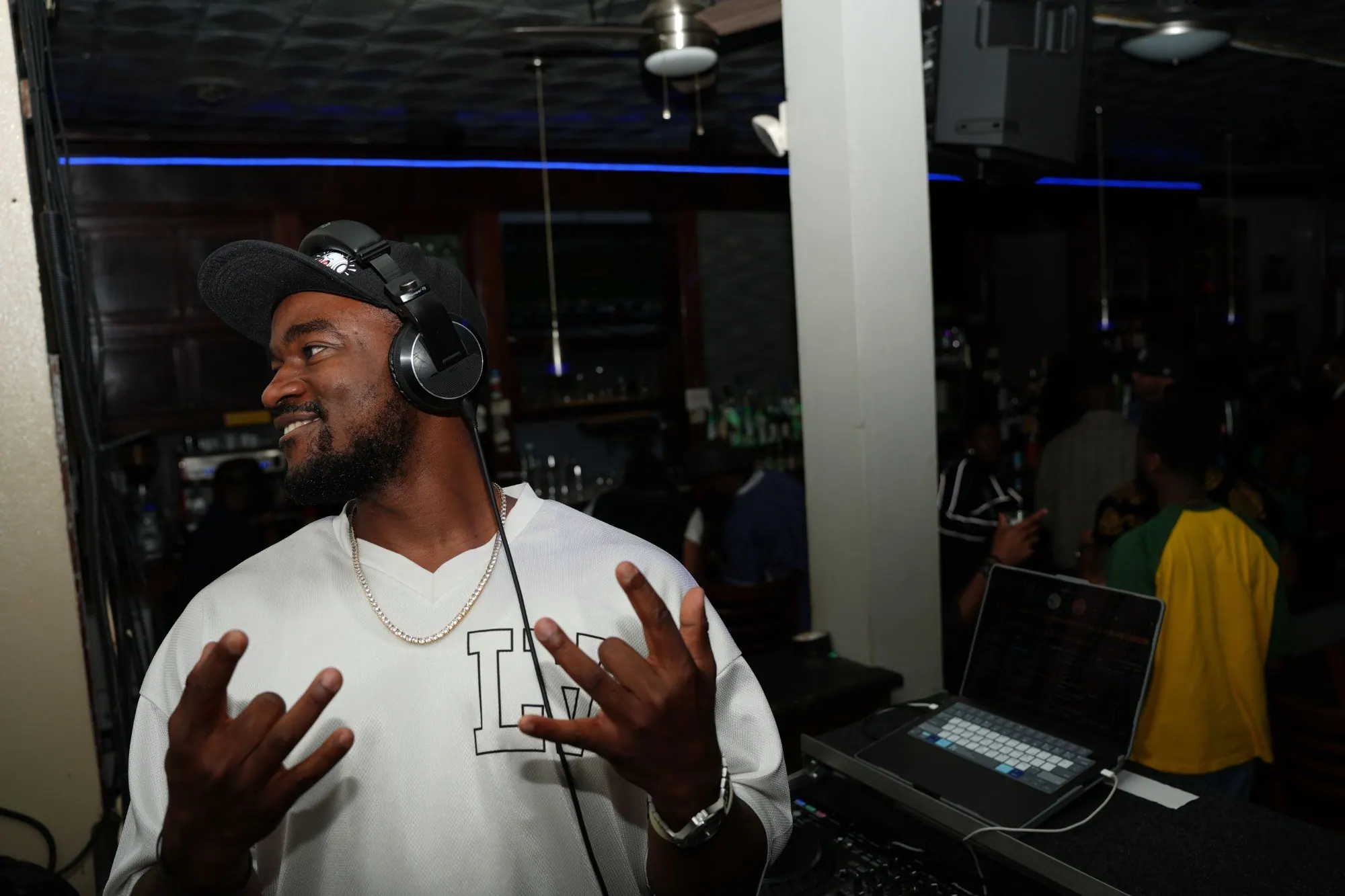 DJ Pharoh mid-set in a bar, headphones on, laughing with his hands up, the crowd behind him.