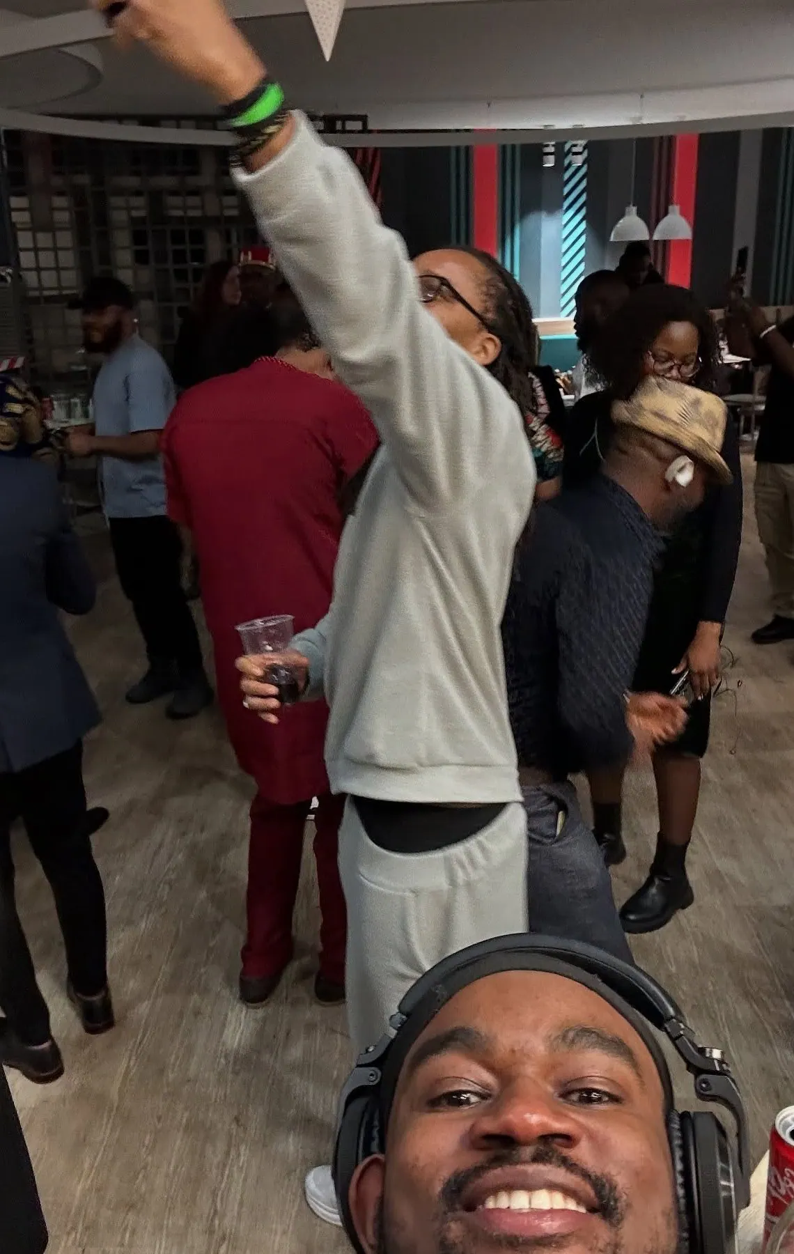 DJ Pharoh in headphones grinning into the camera at a party in Budapest, guests dancing behind him, one with a fist in the air.