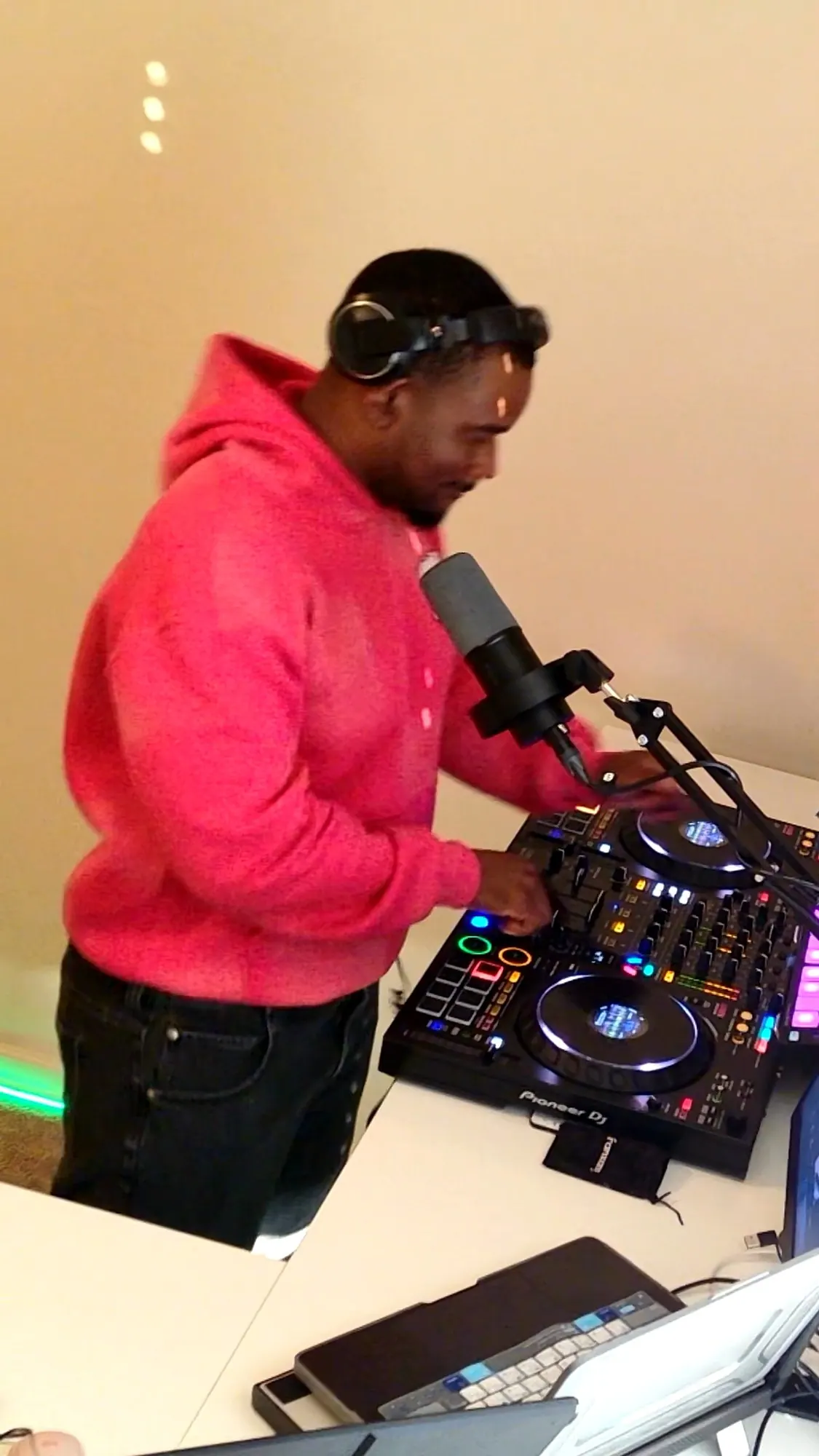DJ Pharoh in a pink hoodie and headphones mixing on a Pioneer controller, a microphone on a boom arm in front of him.