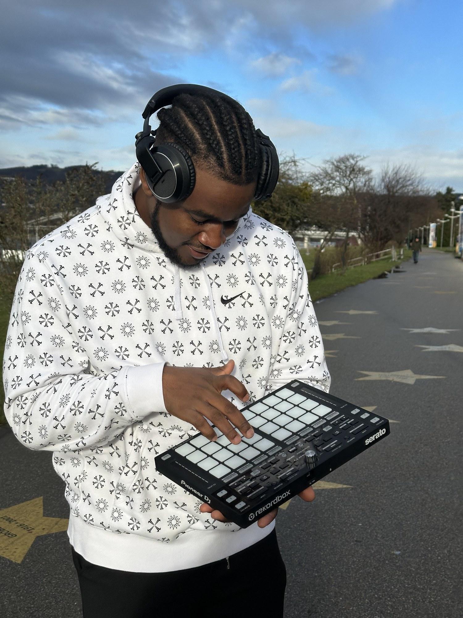 DJ Pharoh in headphones under a wide blue sky, playing a pad controller held in one hand.