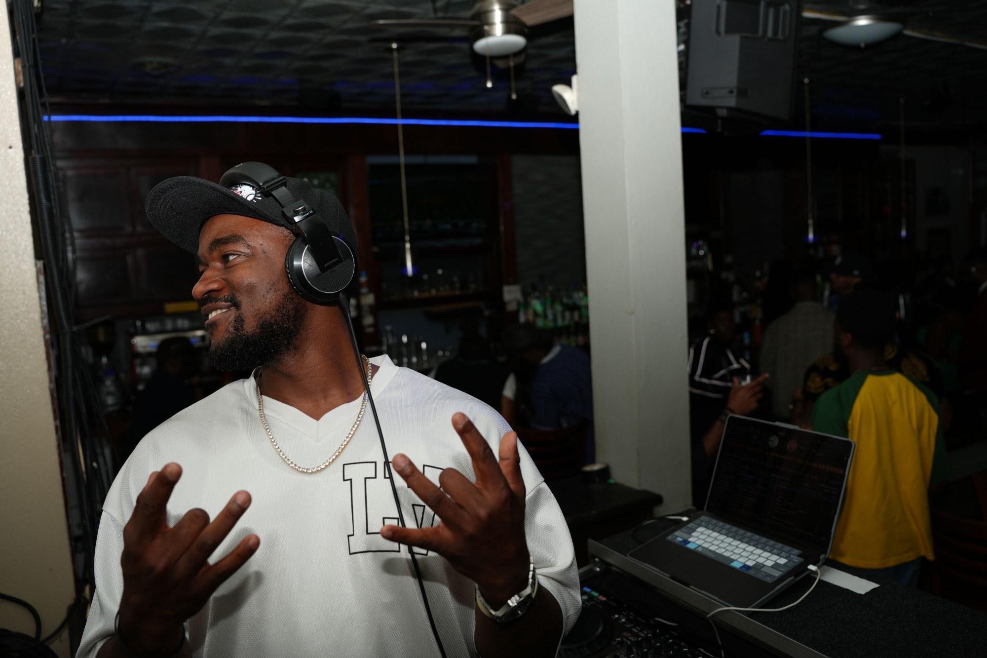 DJ Pharoh mid-set in a bar, headphones on, laughing with his hands up, the crowd behind him.