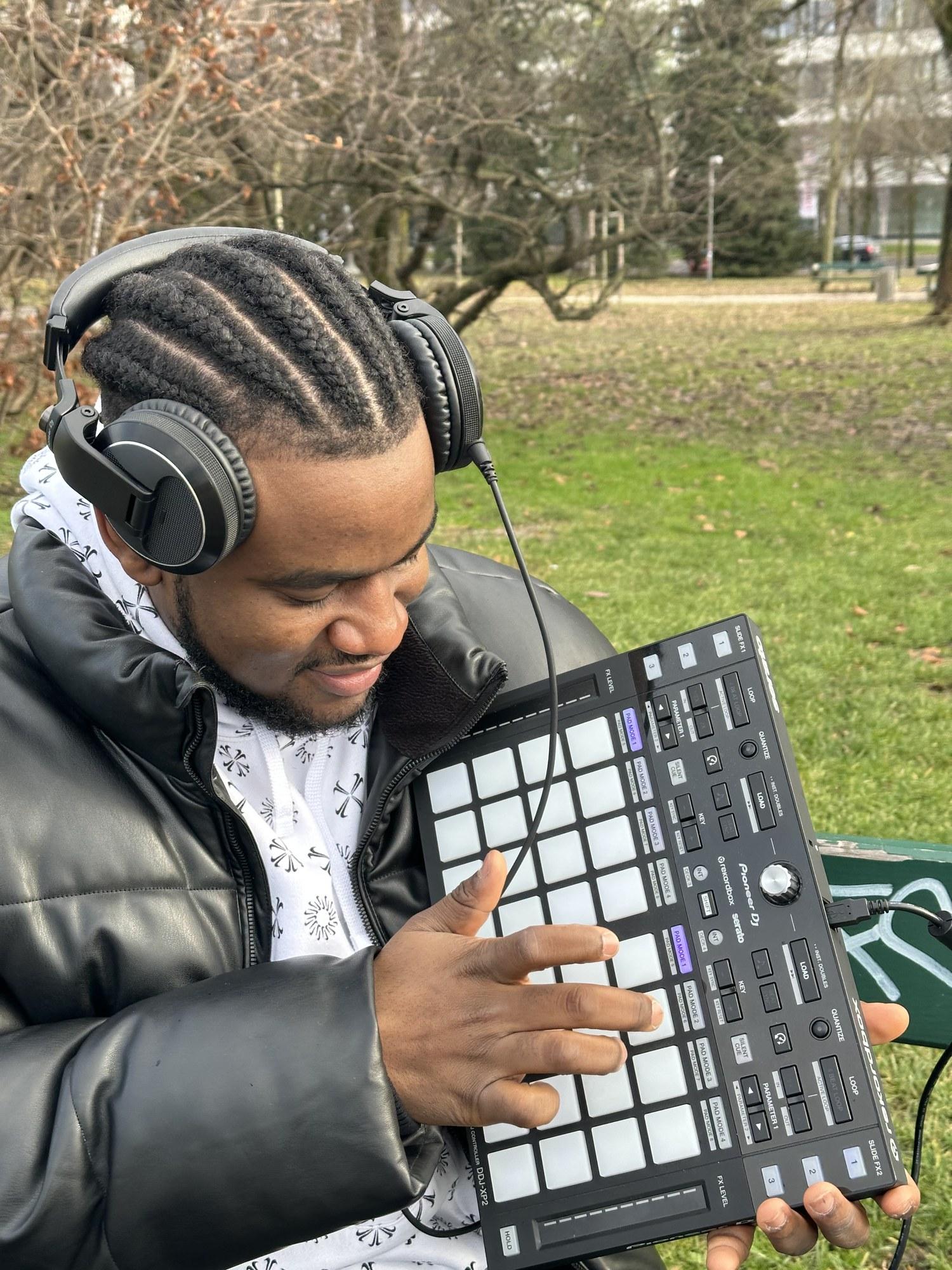 Close on DJ Pharoh in headphones on a park bench, finger drumming on a pad controller.