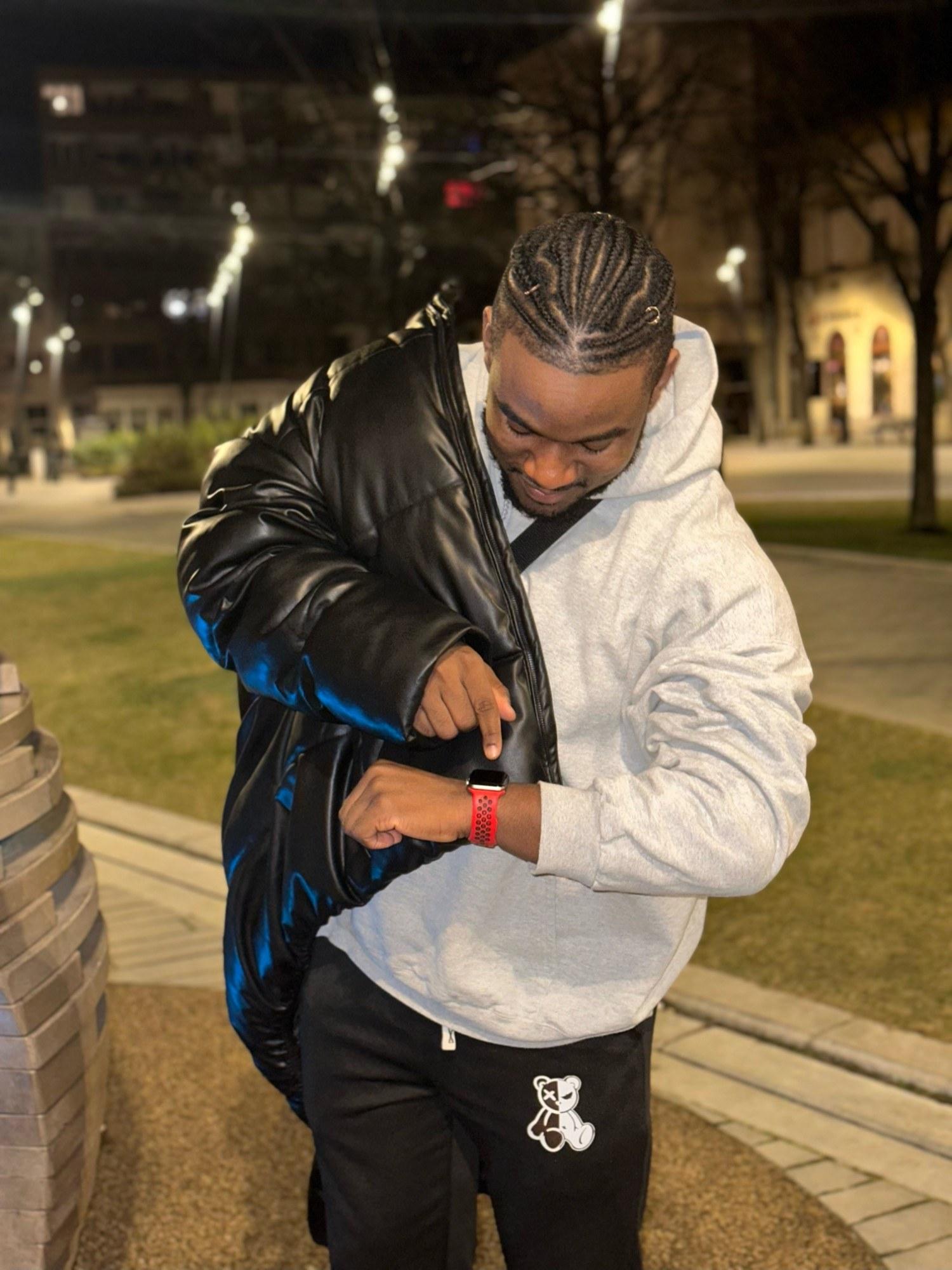 DJ Pharoh at night in a grey hoodie and leather puffer jacket, checking his watch.