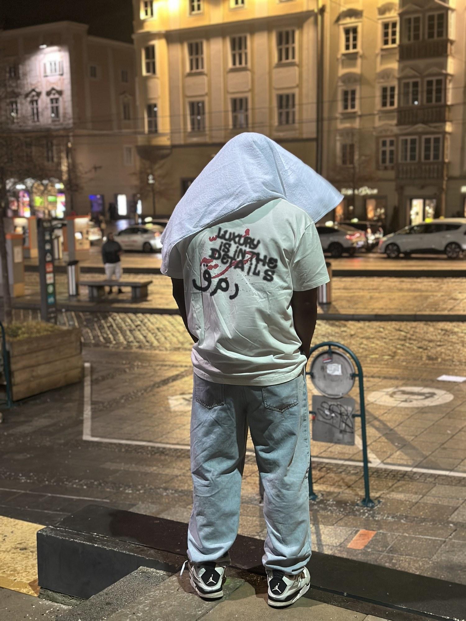 DJ Pharoh from behind in a city square at night. His shirt reads: Luxury is in the details.