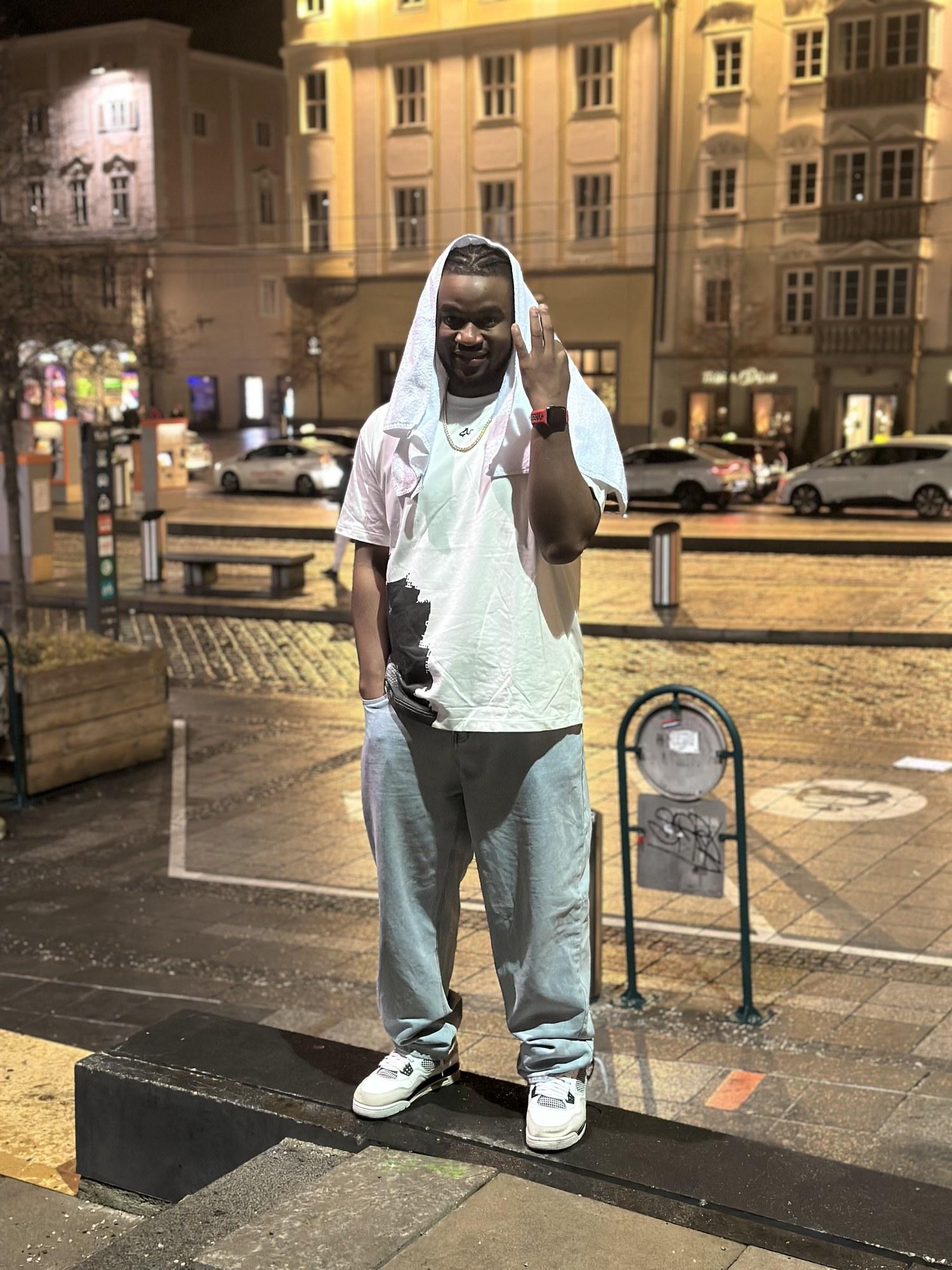 DJ Pharoh grinning in a city square at night, a white towel draped over his head.