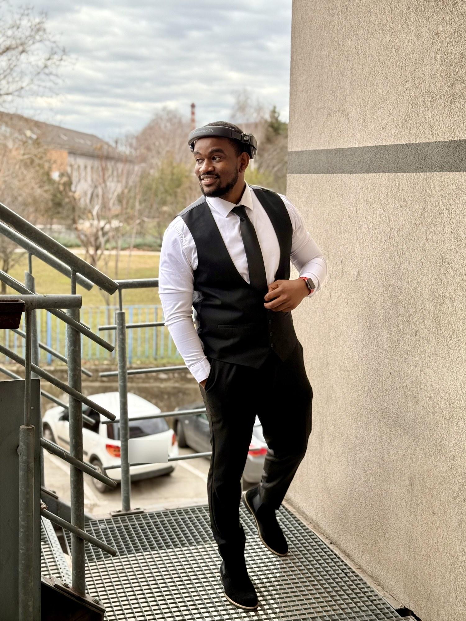 DJ Pharoh in a waistcoat and tie on an outdoor stairway, DJ headphones pushed up on his forehead.