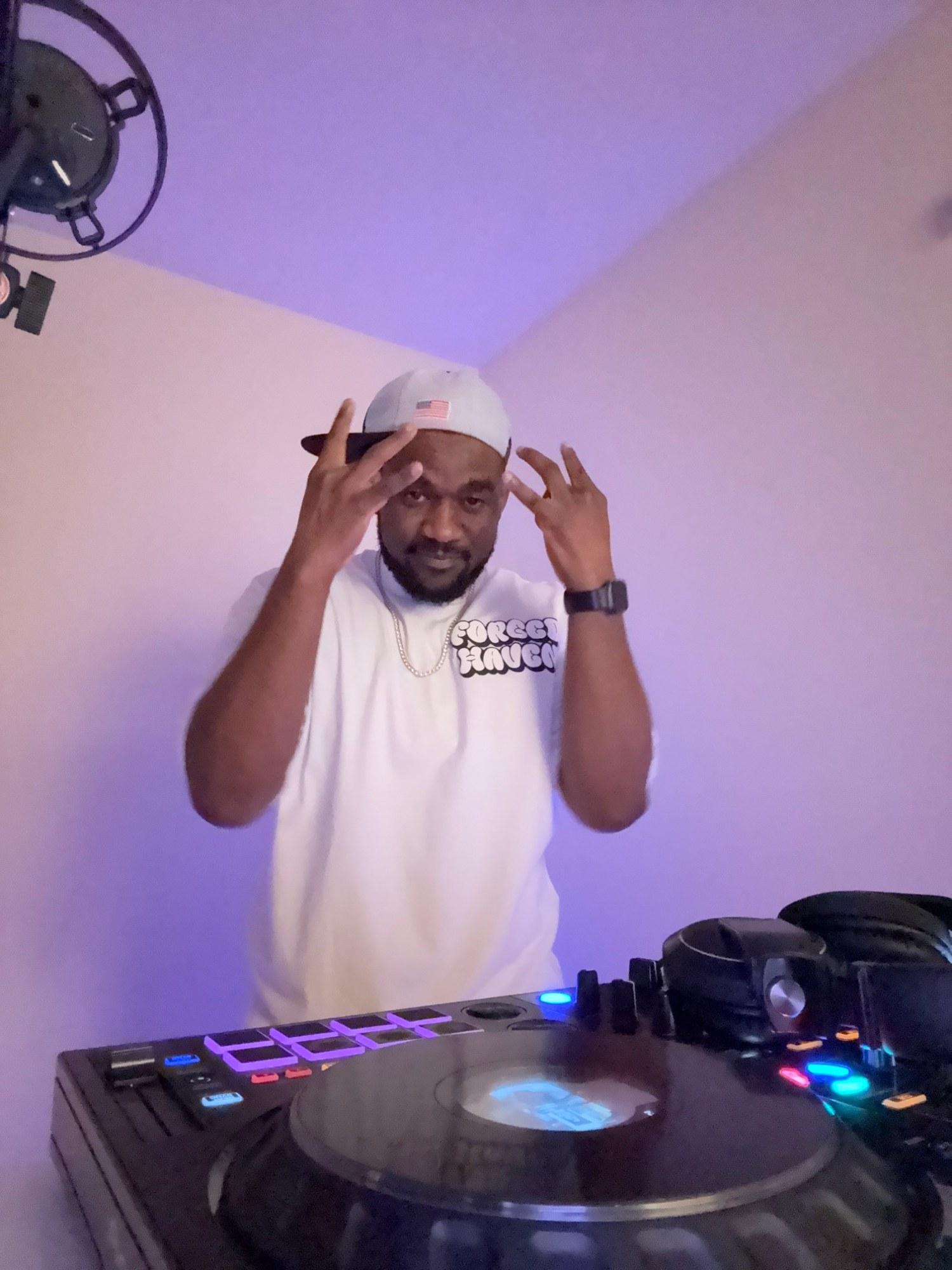 DJ Pharoh behind the decks in a studio washed in violet light, both hands up at his cap.