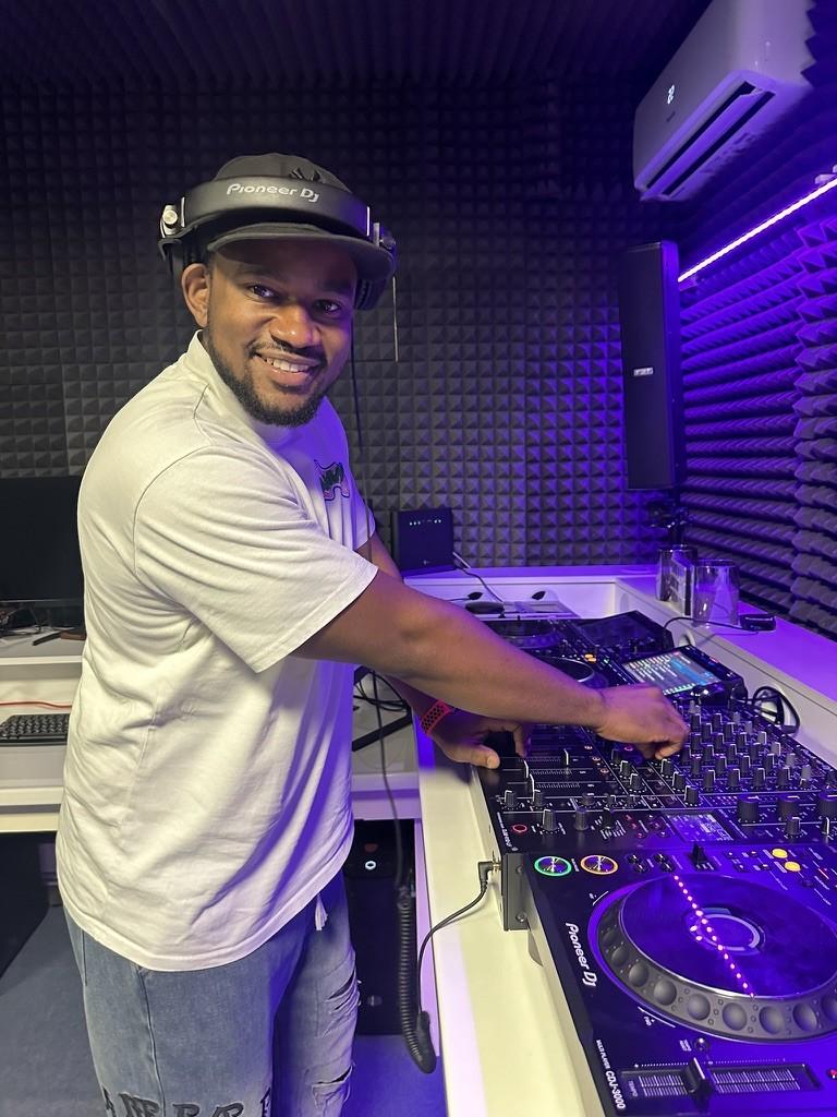 DJ Pharoh smiling behind a Pioneer DJ controller in a studio lit purple, headphones on.